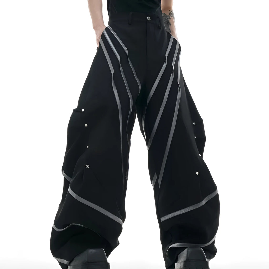 Men's black trousers with three-dimensional cut, metal striped rivet details and pleats on a white background.