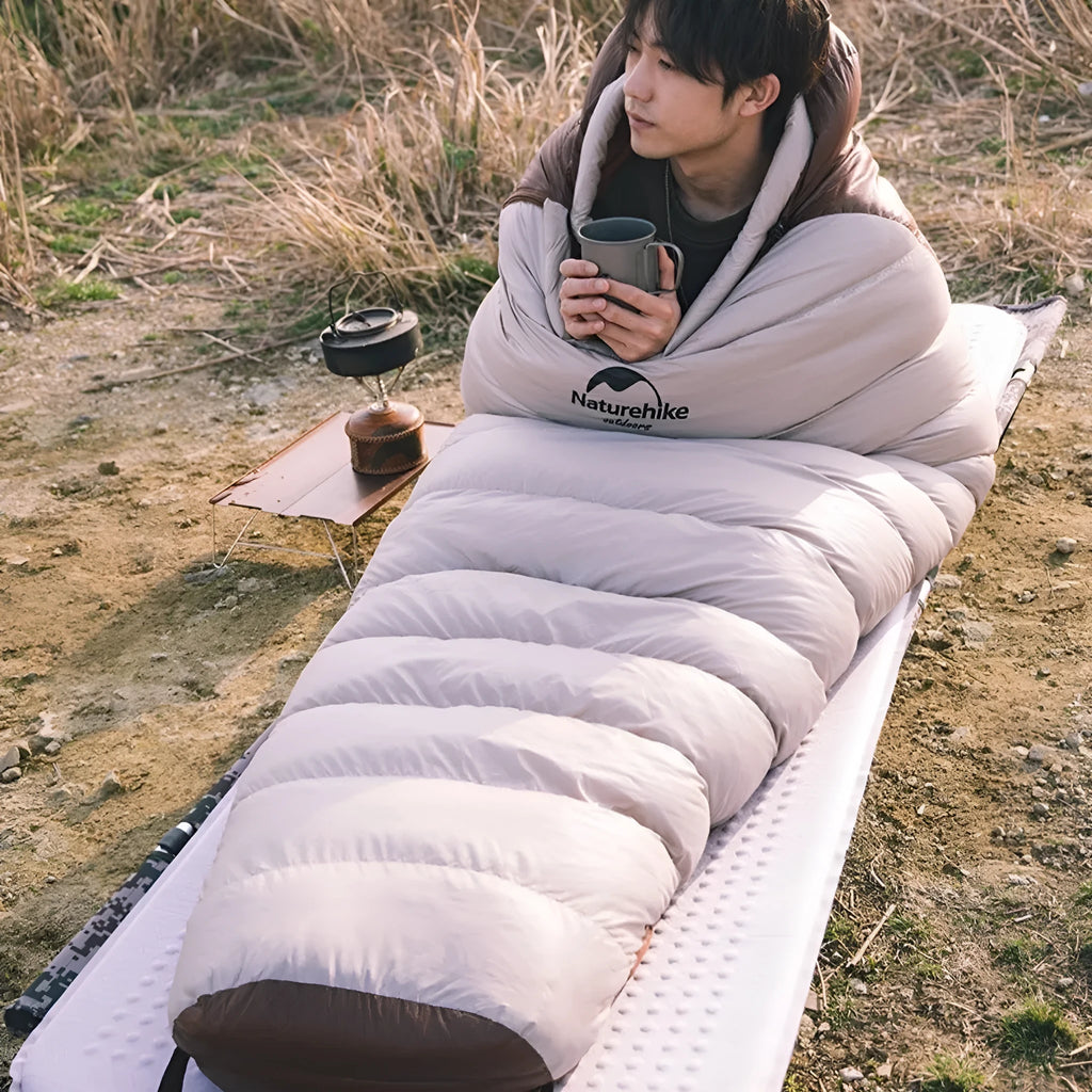 Adult mummy-style goose down sleeping bag in its storage bag, shown in a camping setting.