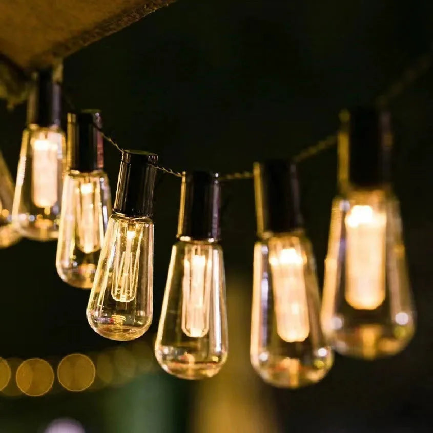 Retro infusion bottle shaped glass bulb outdoor fairy lights with green cable, shown in garden setting.