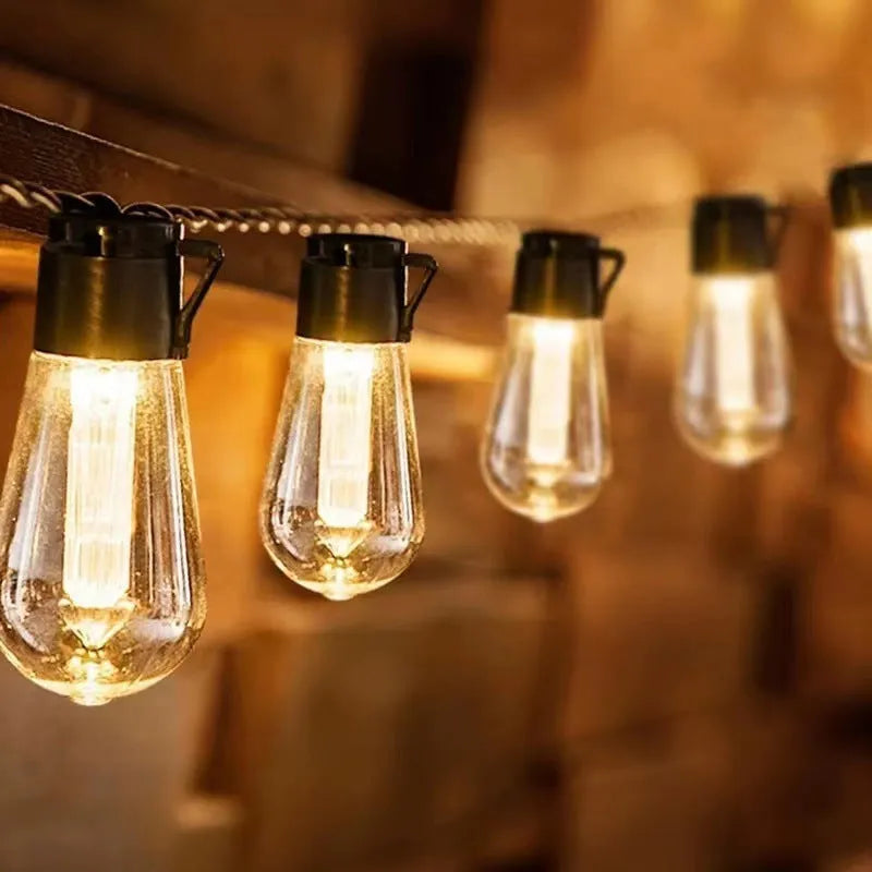 Retro infusion bottle shaped glass bulb outdoor fairy lights with green cable, shown in garden setting.