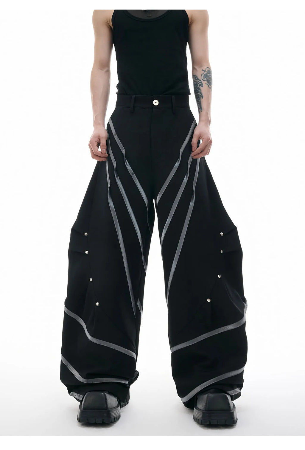 Men's black trousers with three-dimensional cut, metal striped rivet details and pleats on a white background.