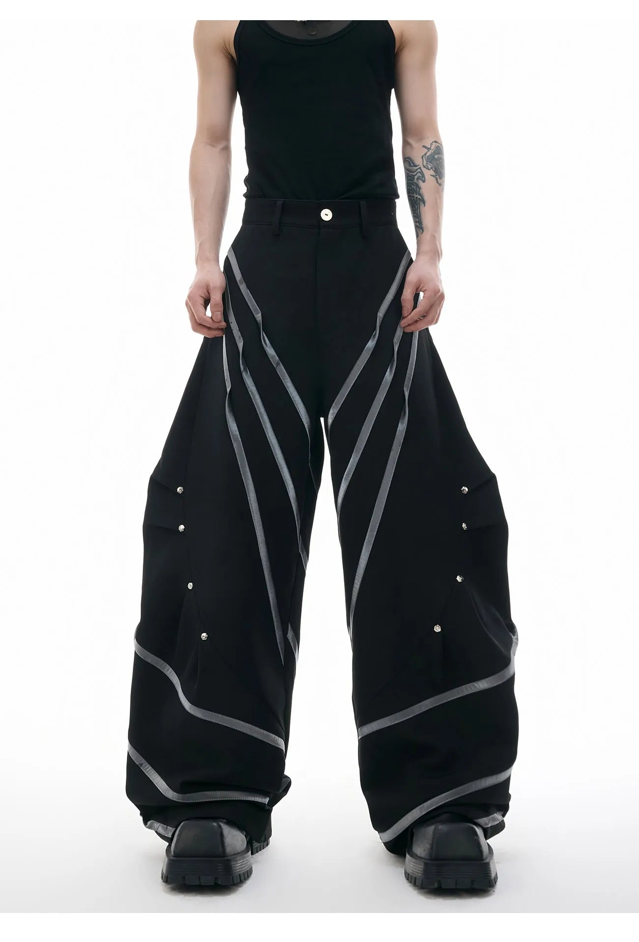 Men's black trousers with three-dimensional cut, metal striped rivet details and pleats on a white background.