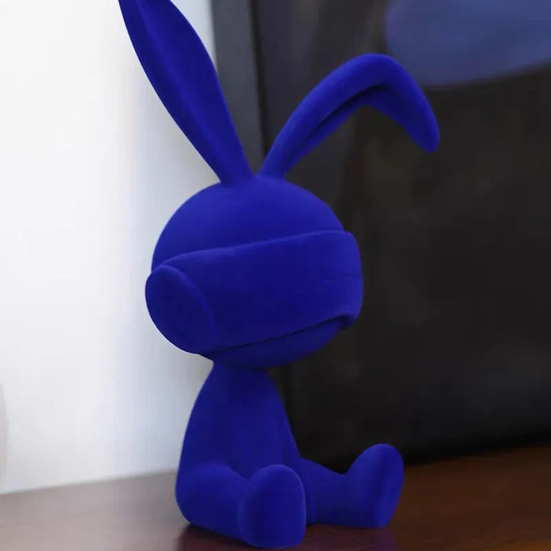 Suede blindfold rabbit resin ornament in green, blue, or red colours on a shelf.