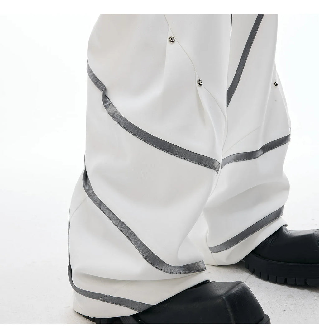 Men's black trousers with three-dimensional cut, metal striped rivet details and pleats on a white background.
